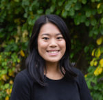 Asian woman standing in front of hedge smiling at the camera, wearing black t-shirt.