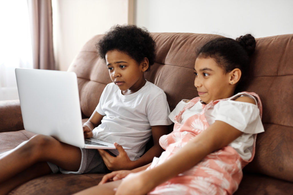 two children sit on a brown couch with a laptop