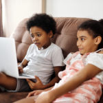 two children sit on a brown couch with a laptop