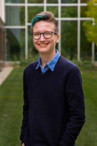 Quinn wearing a blue sweater, standing in front of a glass building on the grass.