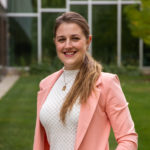 Woman with a ponytail off the right side wearing a pink blazer over a white shirt and necklace. Posture is towards the camera and smiling.