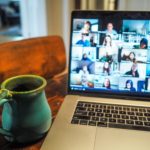 Zoom call on laptop screen placed beside a teal mug on a wooden desk.