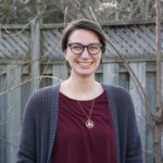 White woman wearing maroon shirt and grey cardigan smiling at camera standing outdoors in front of two trees 