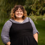 White woman with short brown curly hair wearing black dress overtop blue blouse