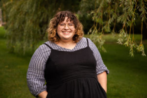 White woman with short brown curly hair wearing black dress overtop blue blouse